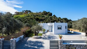 A white building with a stone wall, a paved pathway, and a view of a hillside. - Amazing Summer Villa in Kolympia (Κολύμπια)