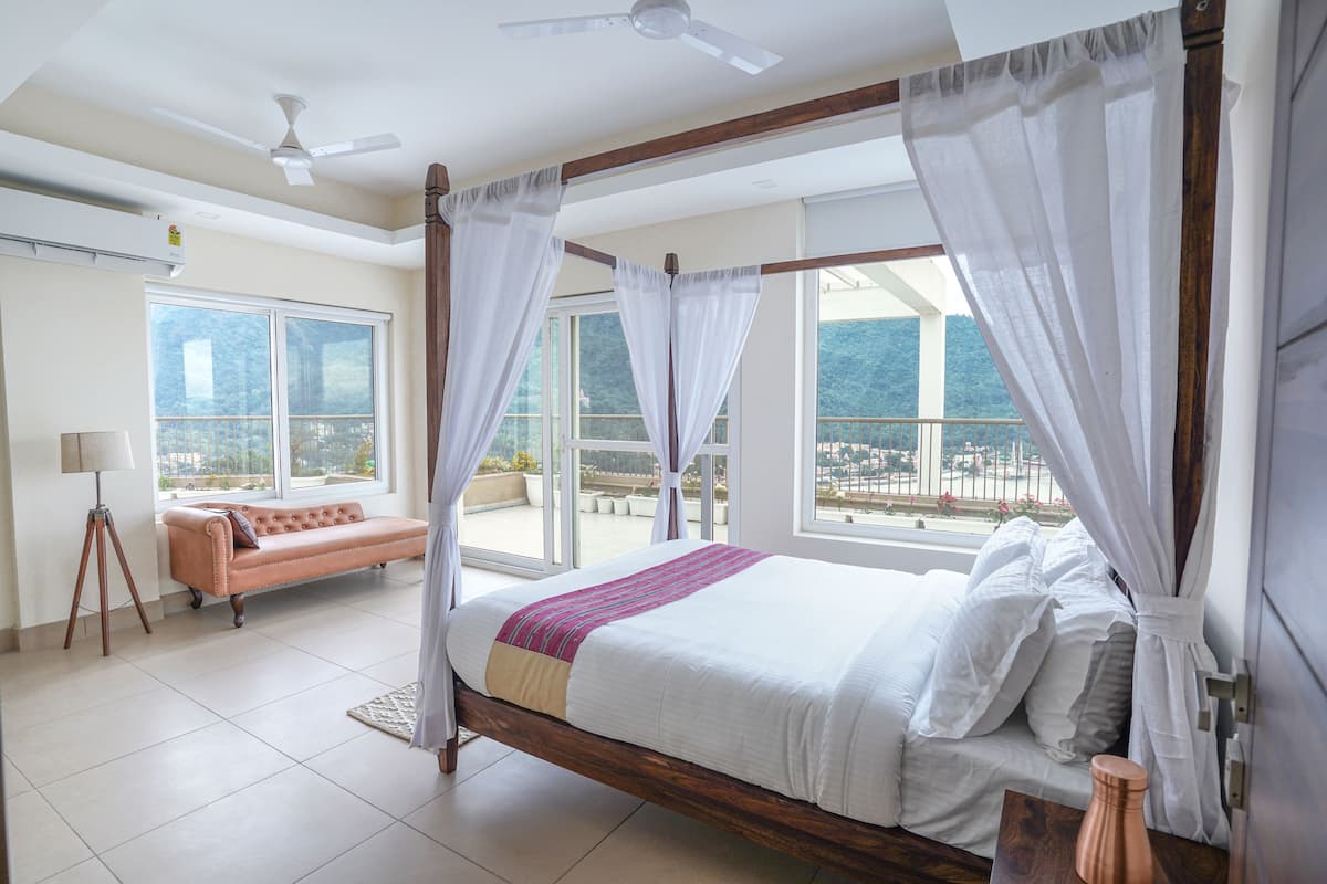 A four-poster bed with white linens, a wooden headboard, and sheer curtains. A chaise lounge is visible in the background, and the room has large windows offering a view of a balcony and greenery.