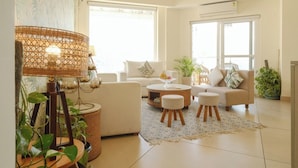 A modern living room with a sofa, coffee table, and ottomans. There are potted plants and a wicker lamp. - Golden Healing Journeys Homestay (Rishikesh)