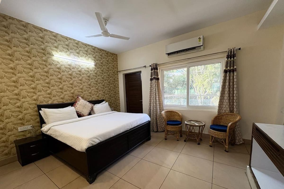 A bedroom with a bed, two wicker chairs, a small table, a ceiling fan, and an air conditioning unit.