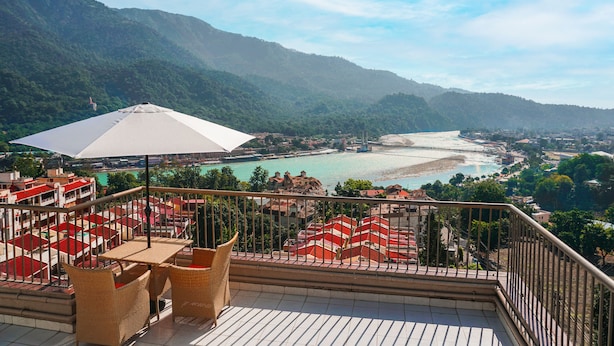 A terrace with wicker furniture and a white umbrella overlooking a coastal town and mountains. - Golden Healing Journeys Homestay (Rishikesh)