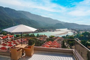 A terrace with wicker furniture and a white umbrella overlooking a coastal town and mountains. - Golden Healing Journeys Homestay (Rishikesh)