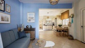 A living room with a sofa, coffee table, and wall art. There is a kitchen in the background with a dining area. - Villa Alysso, fully equipped, modern with shared pool & sea and mountain view (Aptera)