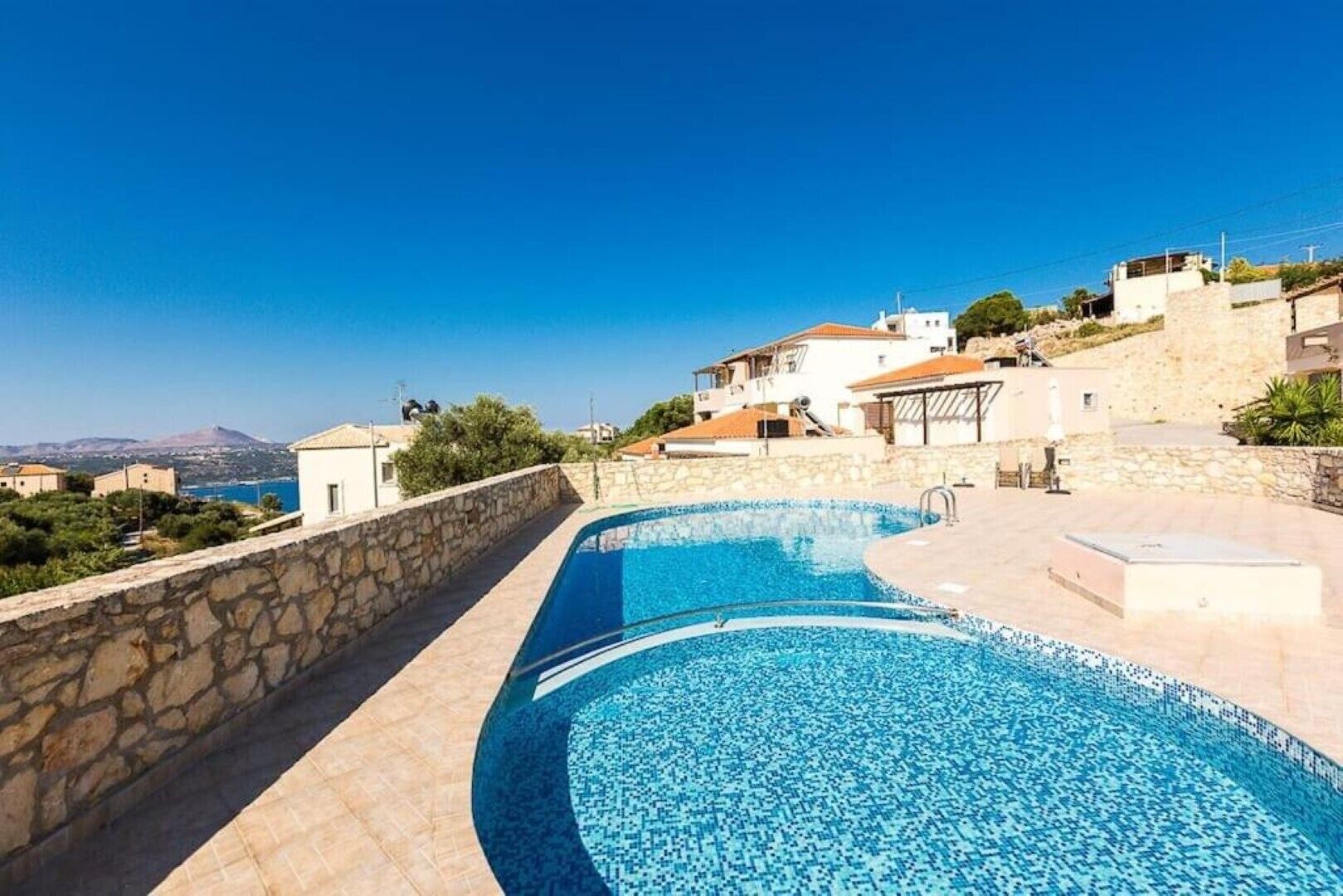 Une piscine bordée d’un mur en pierre, avec vue sur des maisons et une montagne en arrière-plan.
