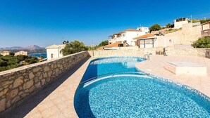 A swimming pool with a stone wall and a view of houses and a mountain in the background. - Villa Alysso, fully equipped, modern with shared pool & sea and mountain view (Aptera)