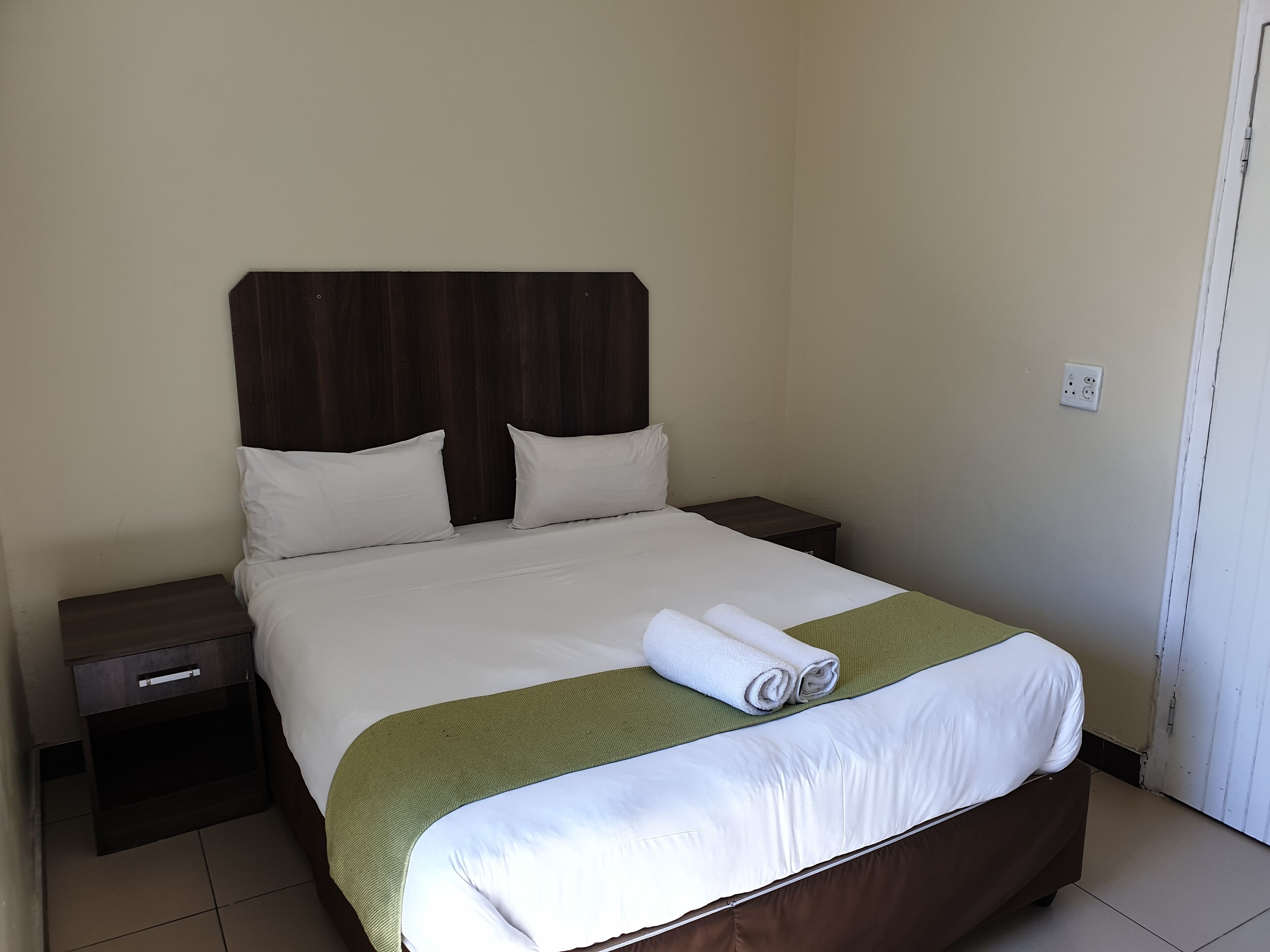 A neatly made bed with white linens and a green accent, a wooden headboard, a small bedside table with a drawer, and a plain wall with a power outlet.