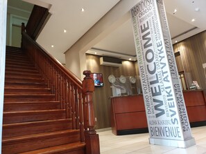 Een hotel receptie met een houten trap, een receptiebalie en een groot welkomstbord.
