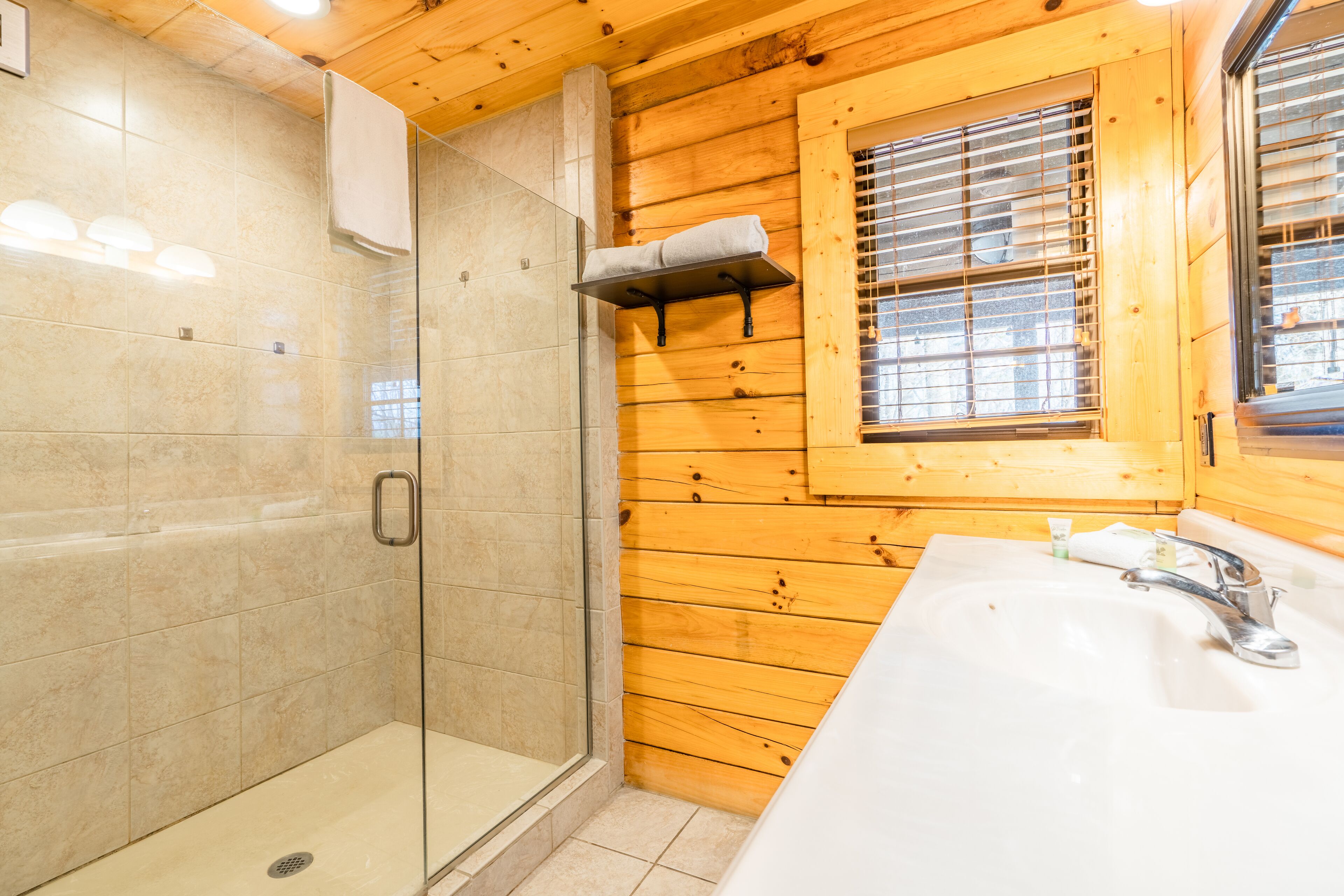 Ein Badezimmer mit einer Duschkabine, einer Holzvertäfelung, einem Fenster mit Jalousien und einem Waschbecken mit Wasserhahn.