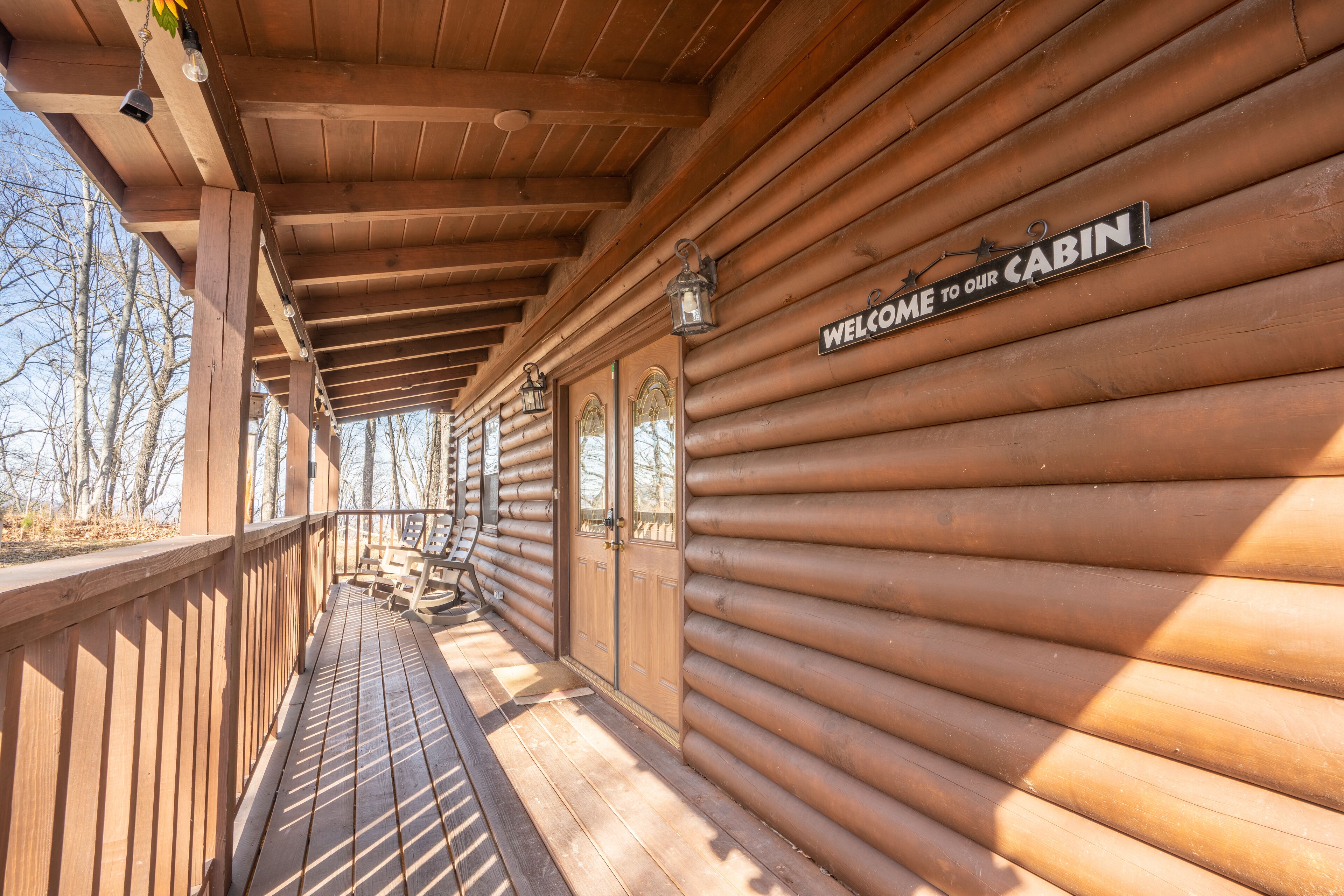A wooden cabin with a covered porch, a welcome sign, and a view of the surrounding trees.