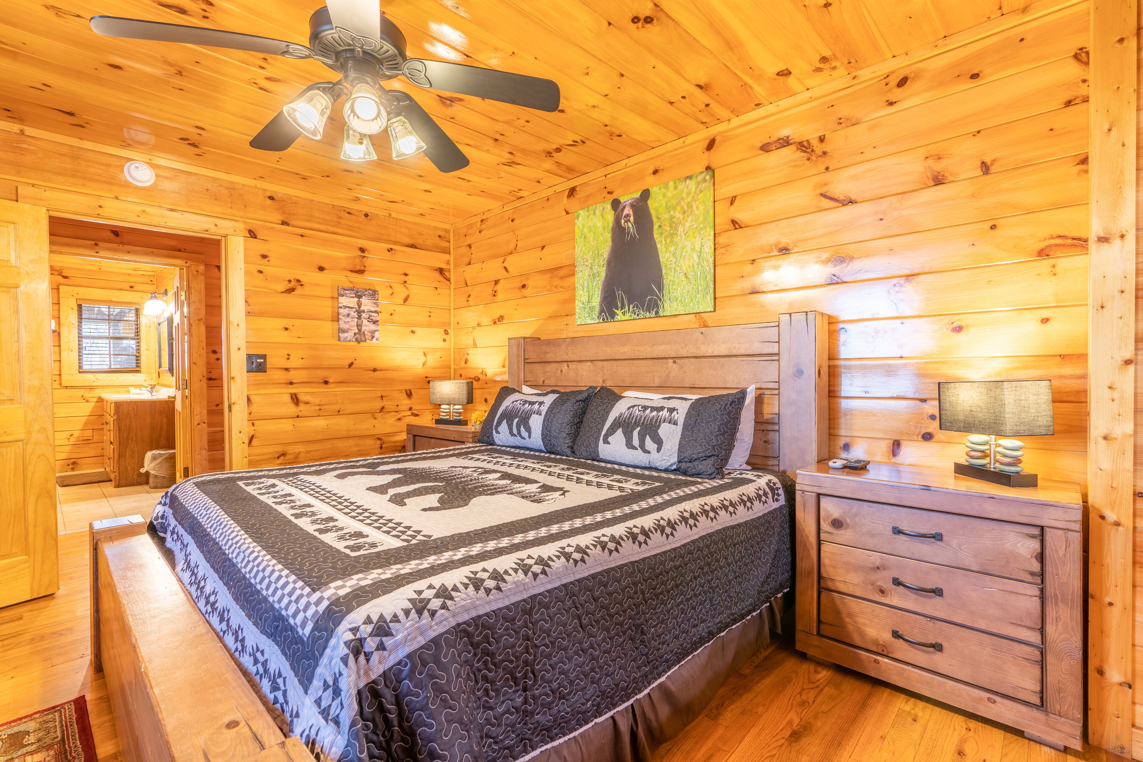 A wooden cabin bedroom with a bed, a nightstand, a dresser, a ceiling fan, and a bear-themed wall decoration.