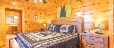 A wooden cabin bedroom with a bed, a nightstand, a dresser, a ceiling fan, and a bear-themed wall decoration.