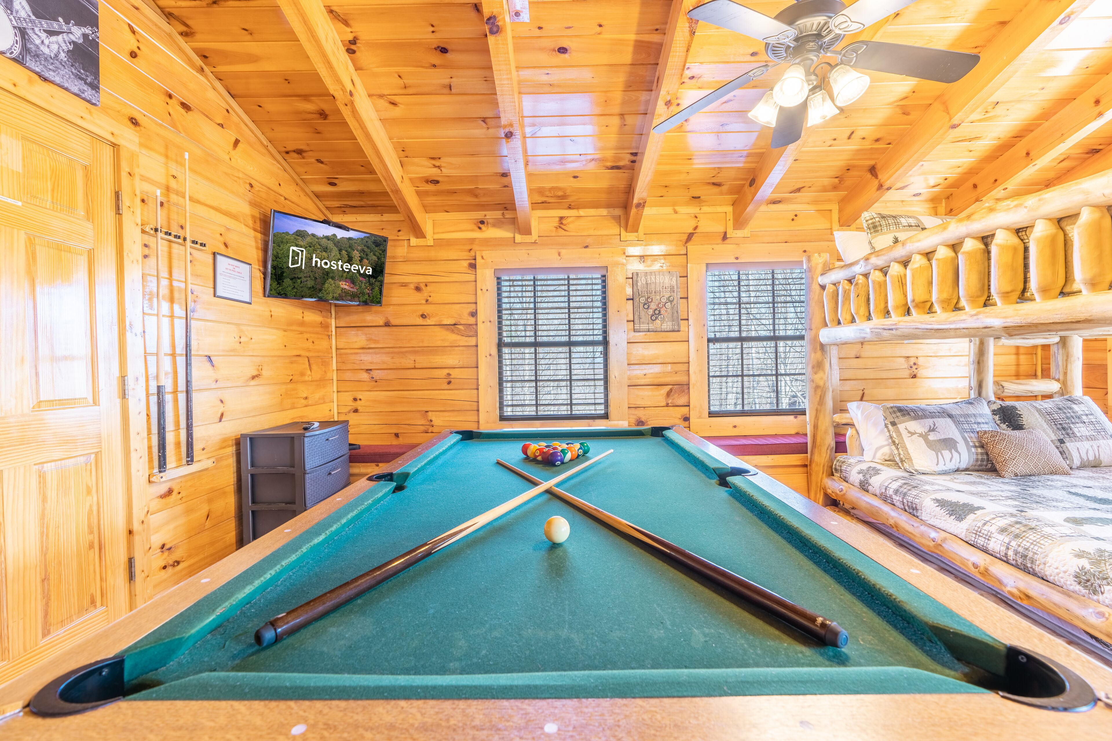 Een pooltafel in een houten chalet met een plafondventilator, een bank met een hertenprint, en een televisie waarop 'hosteeva' staat.