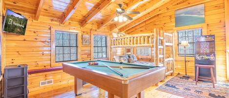 A wooden cabin interior with a pool table, a ceiling fan, and a television displaying 'hosteeva'.