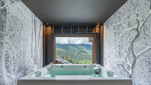 Ein modernes Badezimmer mit einer großzügigen Badewanne, einem Fenster mit Panoramablick auf die Berge und einer strukturierten Wandgestaltung.
