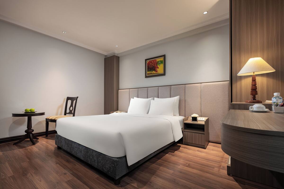 A hotel room with a large bed, bedside tables, a chair, a small table with apples, and a painting on the wall.