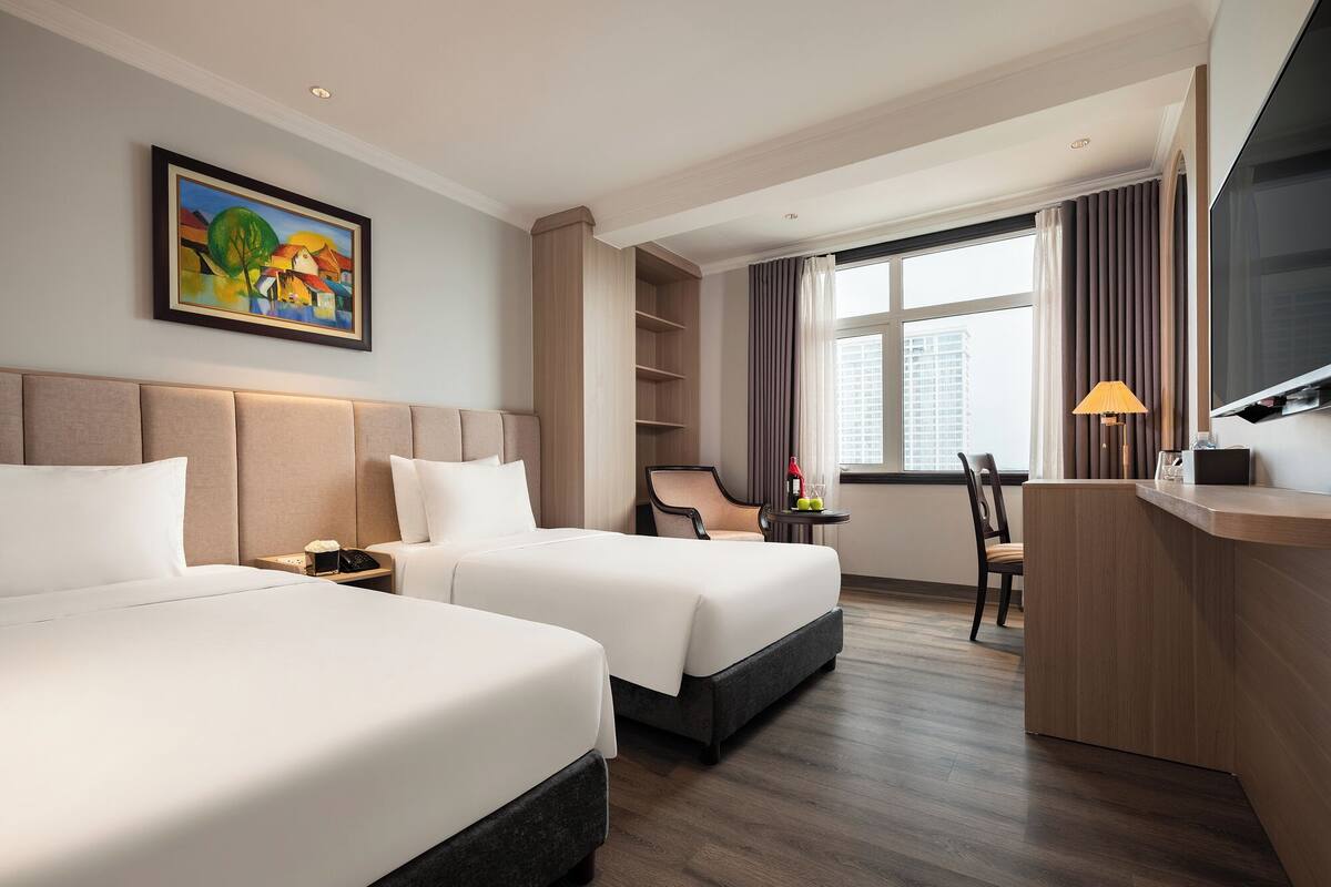 A hotel room with two beds, a desk, a chair, a TV, and a painting on the wall.