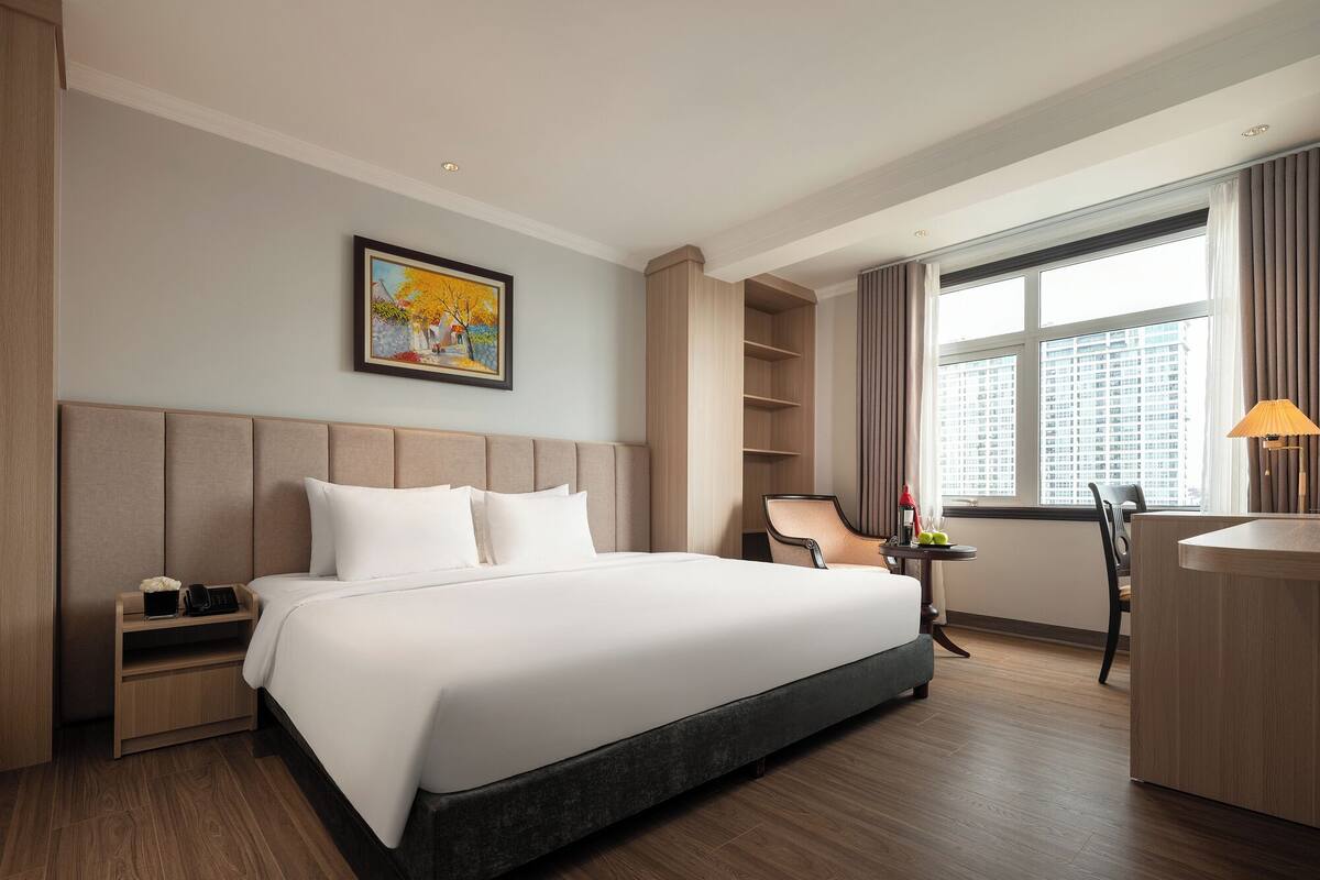 A hotel room with a large bed, a desk, a chair, a small table with a lamp, and a window with curtains.
