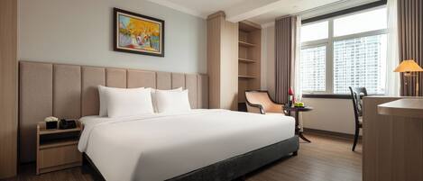 A hotel room with a large bed, a desk, a chair, a small table with a lamp, and a window with curtains.