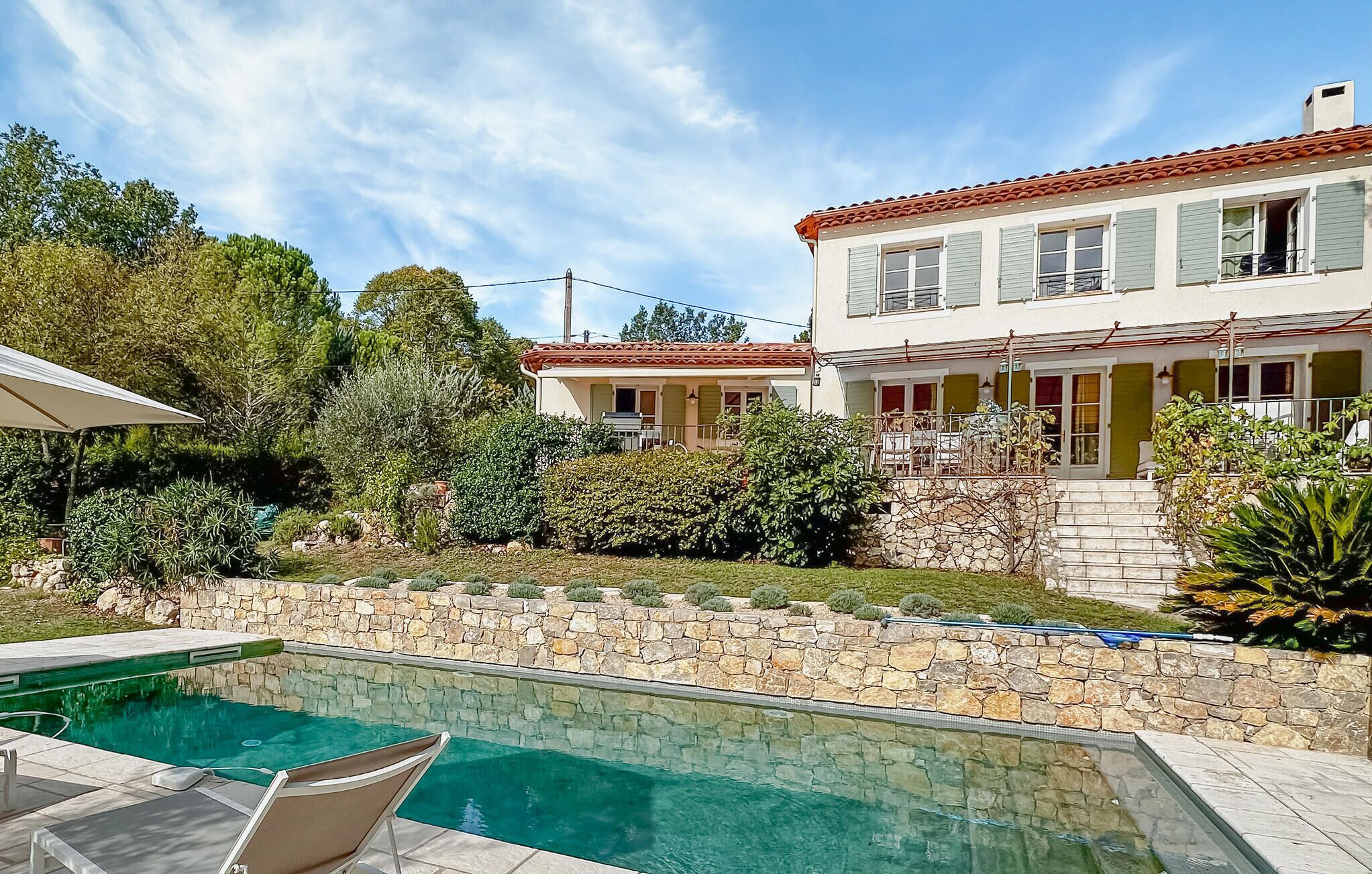 A two-story house with a swimming pool, stone wall, and greenery.