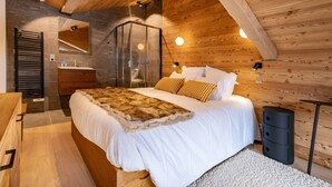 Une chambre avec un plafond en bois, un grand lit avec une literie blanche, un plaid en fourrure et une salle de bain dotée d’une douche à parois de verre.
