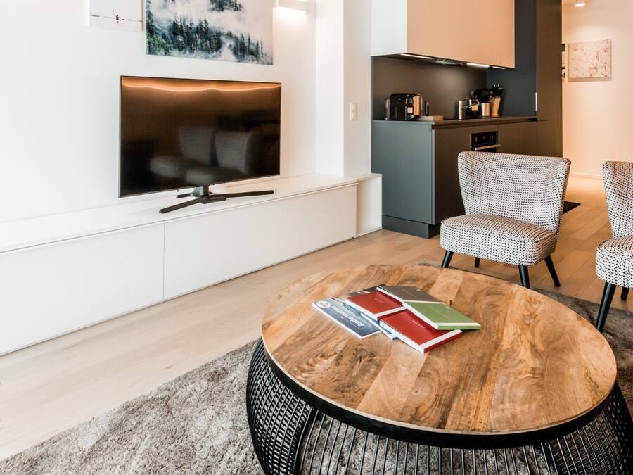 A modern living room with a round coffee table, a flat-screen TV, and a kitchen area with a built-in oven and microwave.
