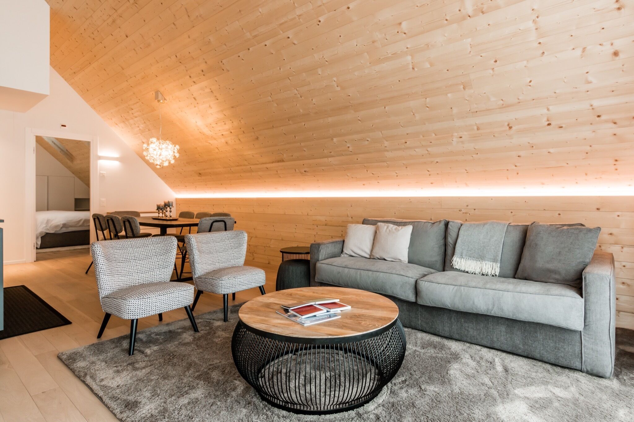 Una sala de estar moderna con techo de madera, sofá gris, sillas estampadas, una mesa de centro redonda y una alfombra.