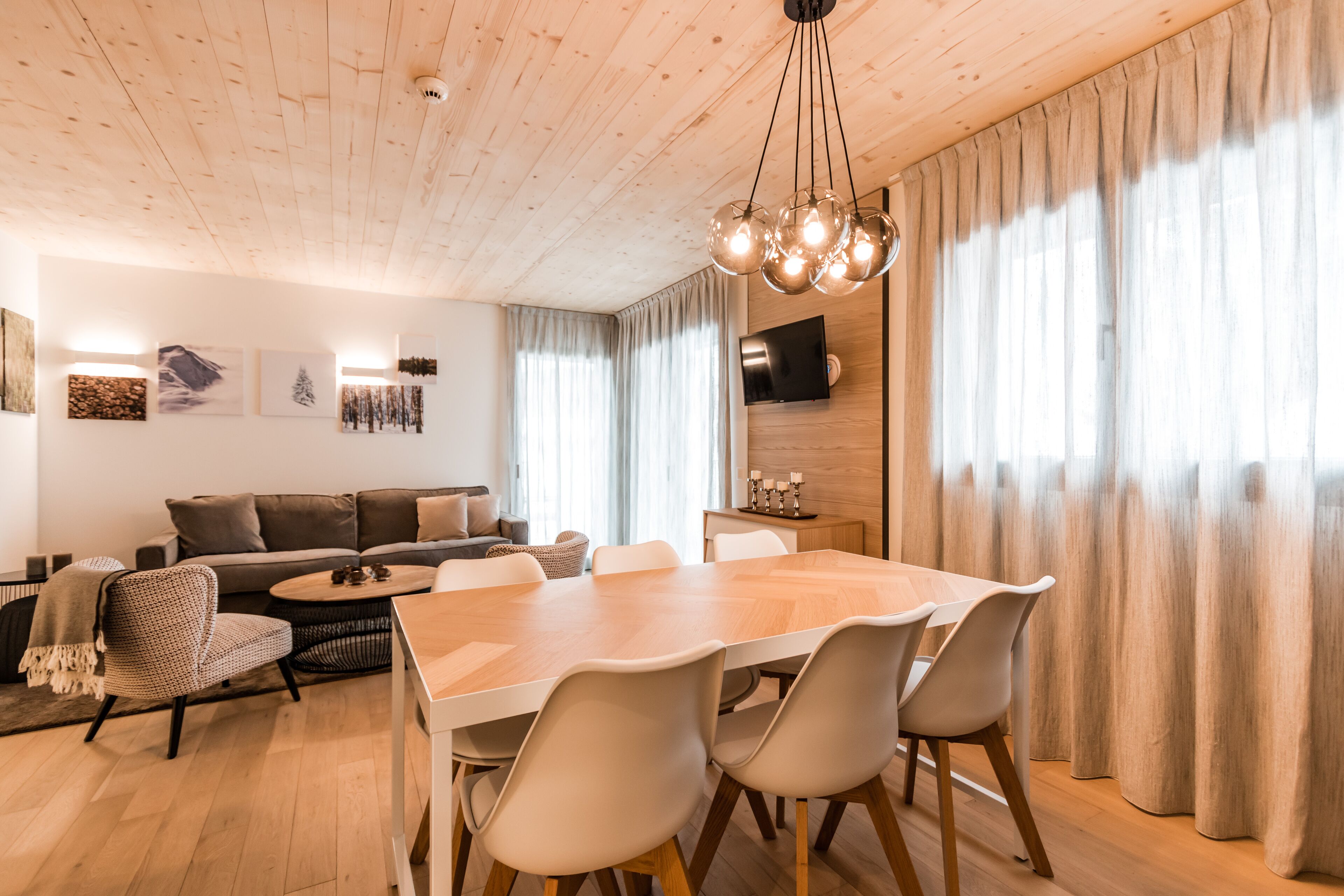 A modern living room with a wooden ceiling, a dining table with chairs, a sofa, and a television mounted on the wall.