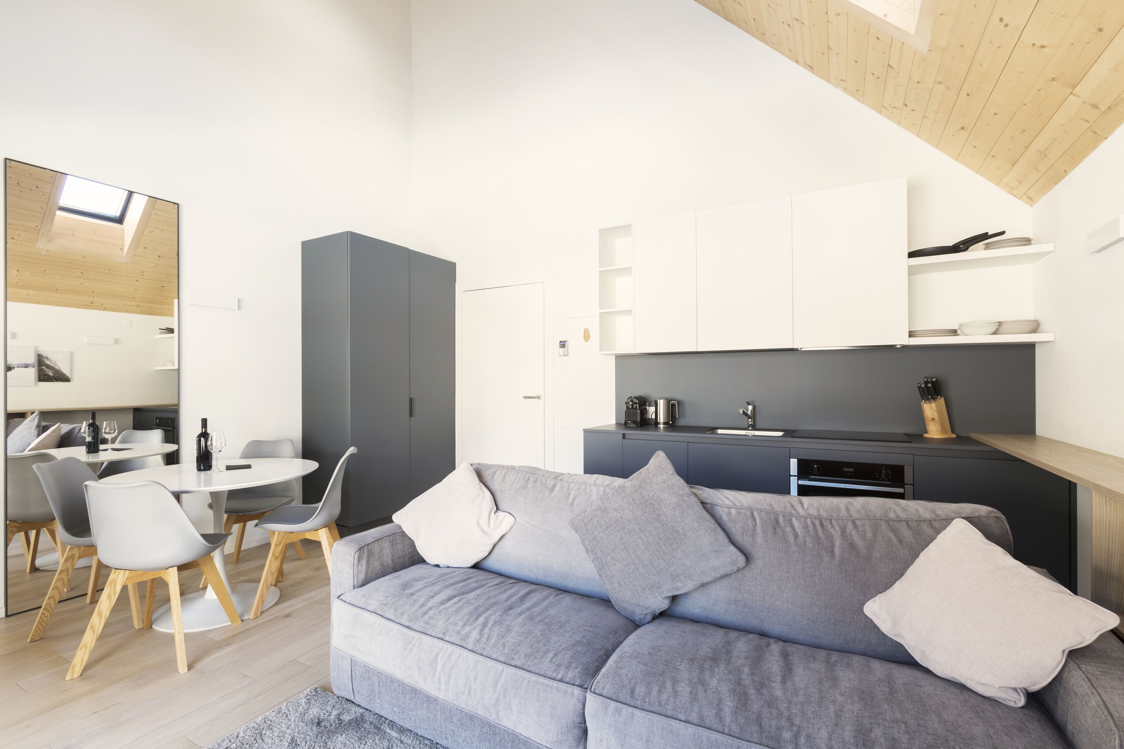 A modern living room with a grey sofa, white cushions, and a dining area with a round table and chairs.