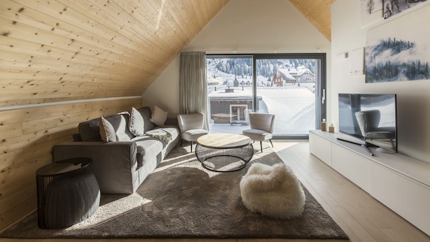 A modern living room with a sloped ceiling, a large window with a snowy view, a sofa set, a round coffee table, and a flat-screen TV. - THE BASE  (Andermatt)