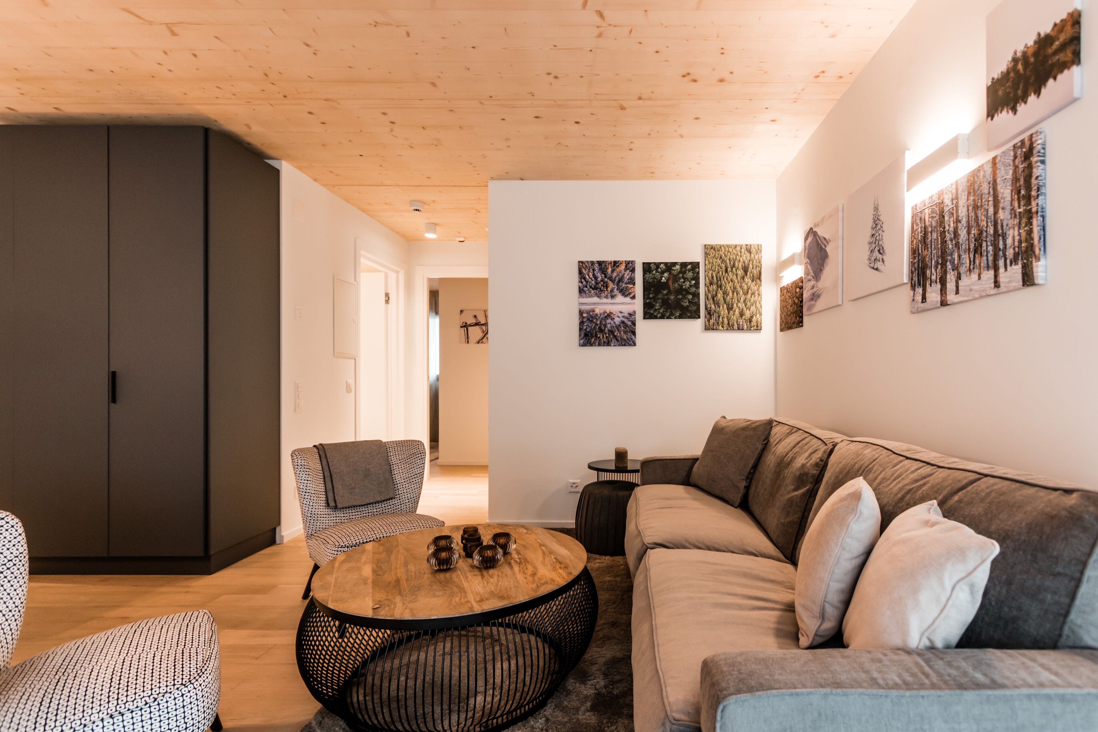 Un salon moderne avec un plafond en bois, un canapé gris, un fauteuil à motifs, une table basse ronde et des œuvres d’art accrochées au mur.