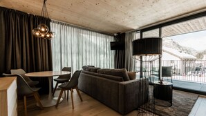 A modern living room with a wooden ceiling, a sofa, a dining table with chairs, a floor lamp, and a view of mountains through large windows. - THE BASE  (Andermatt)