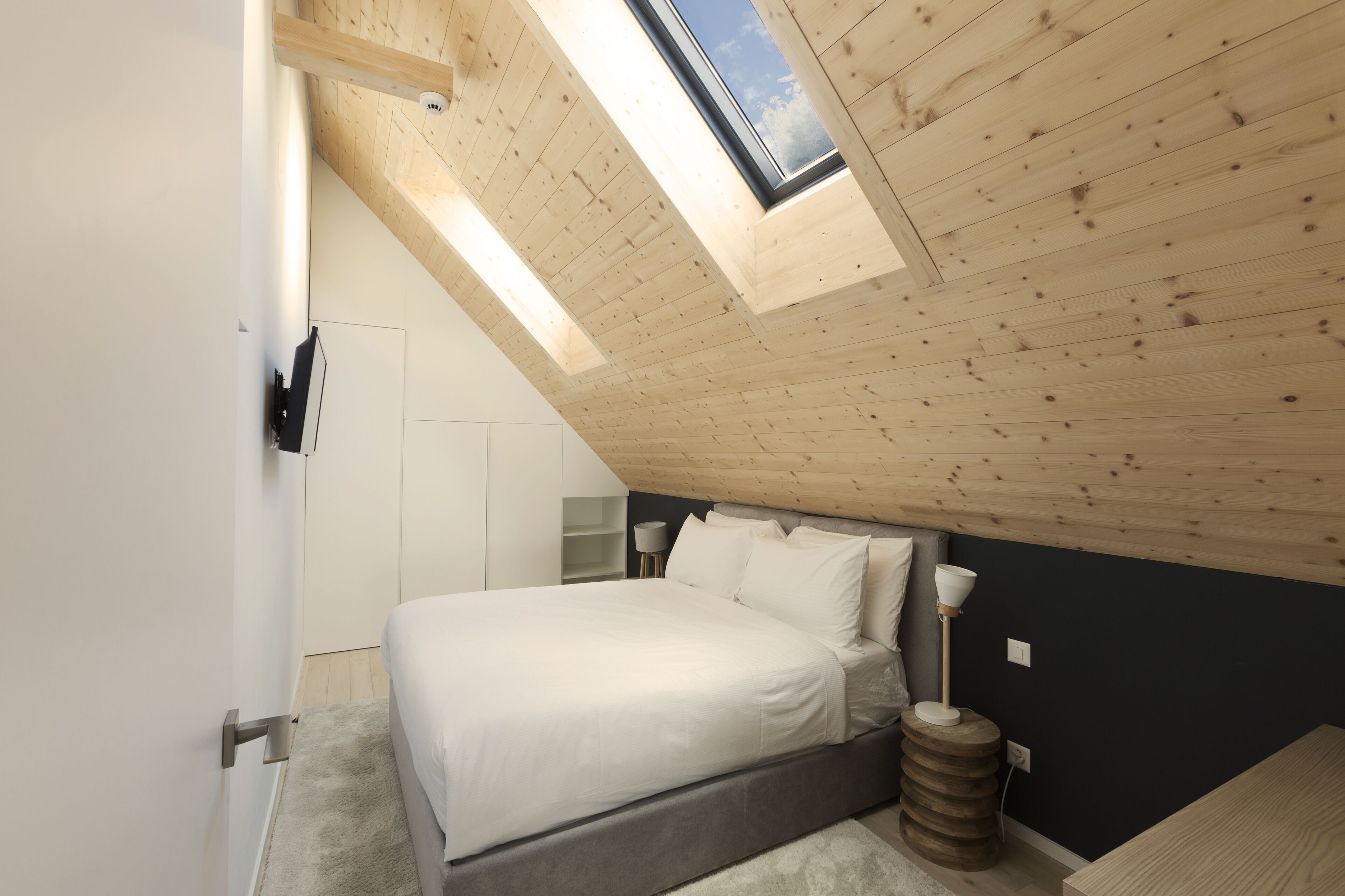 A bedroom with a skylight, a bed, a bedside table, and a lamp.