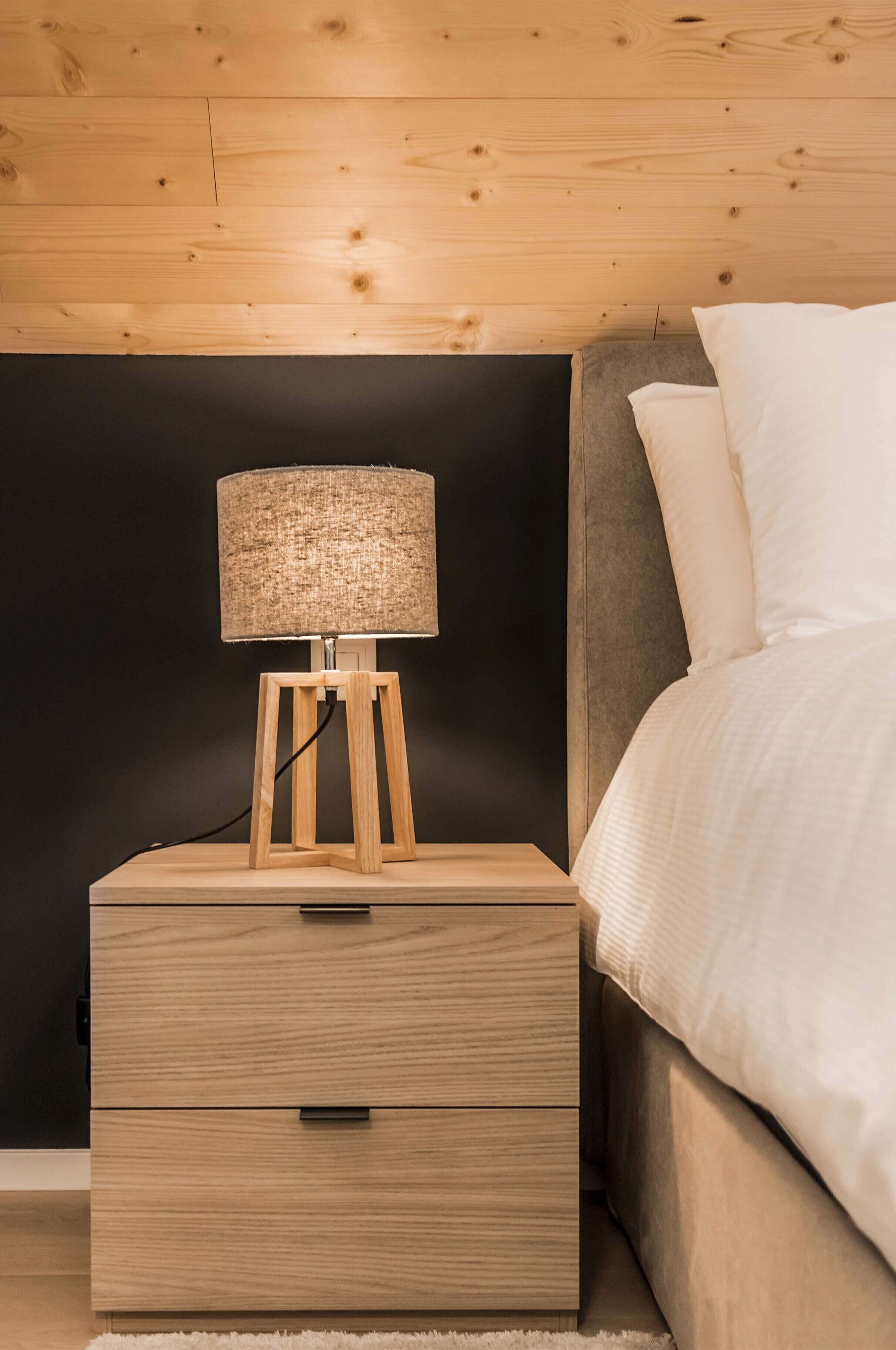 A wooden bedside table with a lamp, a drawer, and a bed with white bedding.