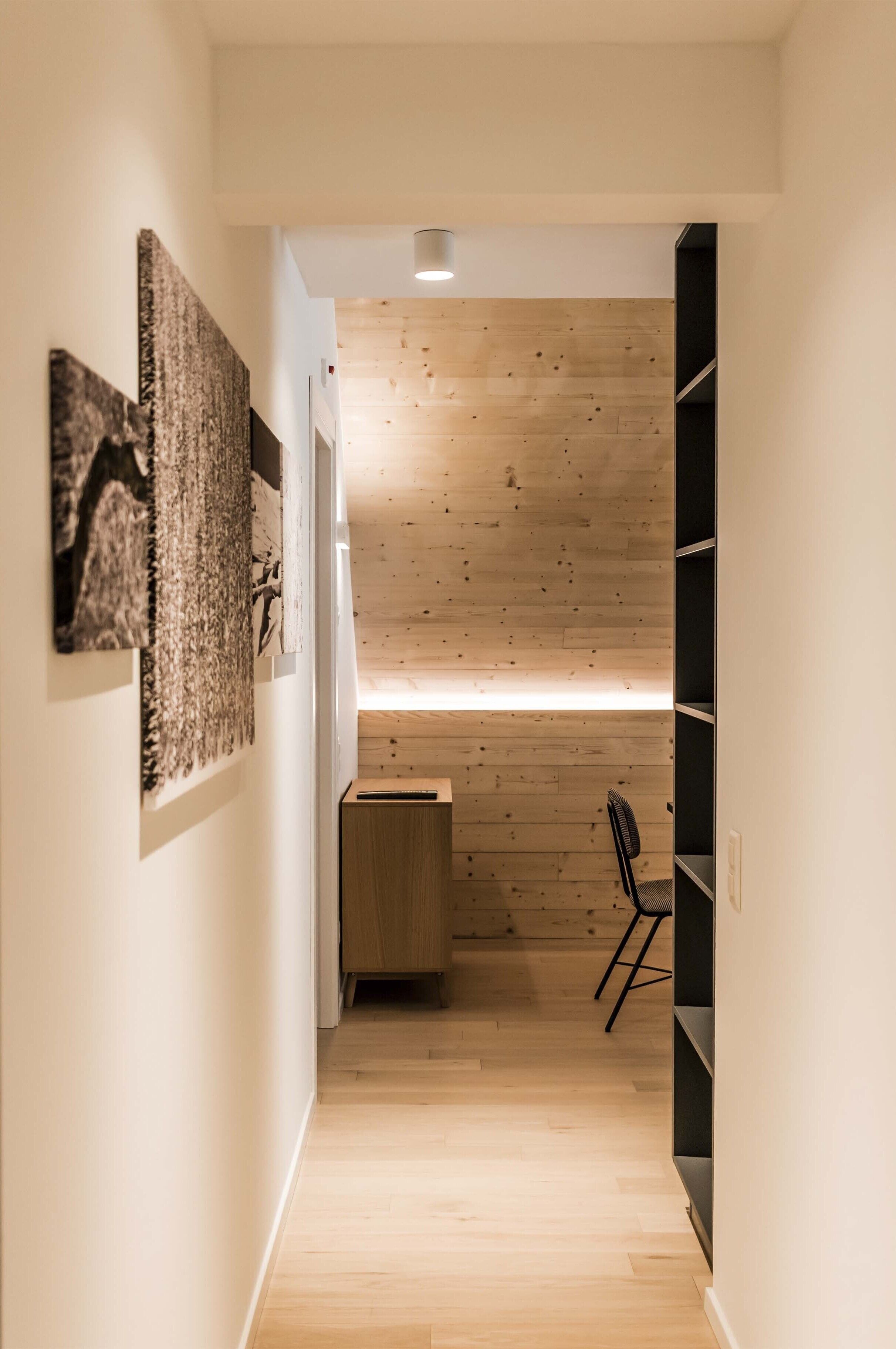 A narrow hallway with wooden flooring, a small desk, a chair, and a black shelving unit.