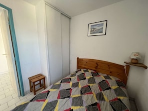 A bedroom with a wooden bed, a small wooden stool, a picture on the wall, and a door with a blue frame.