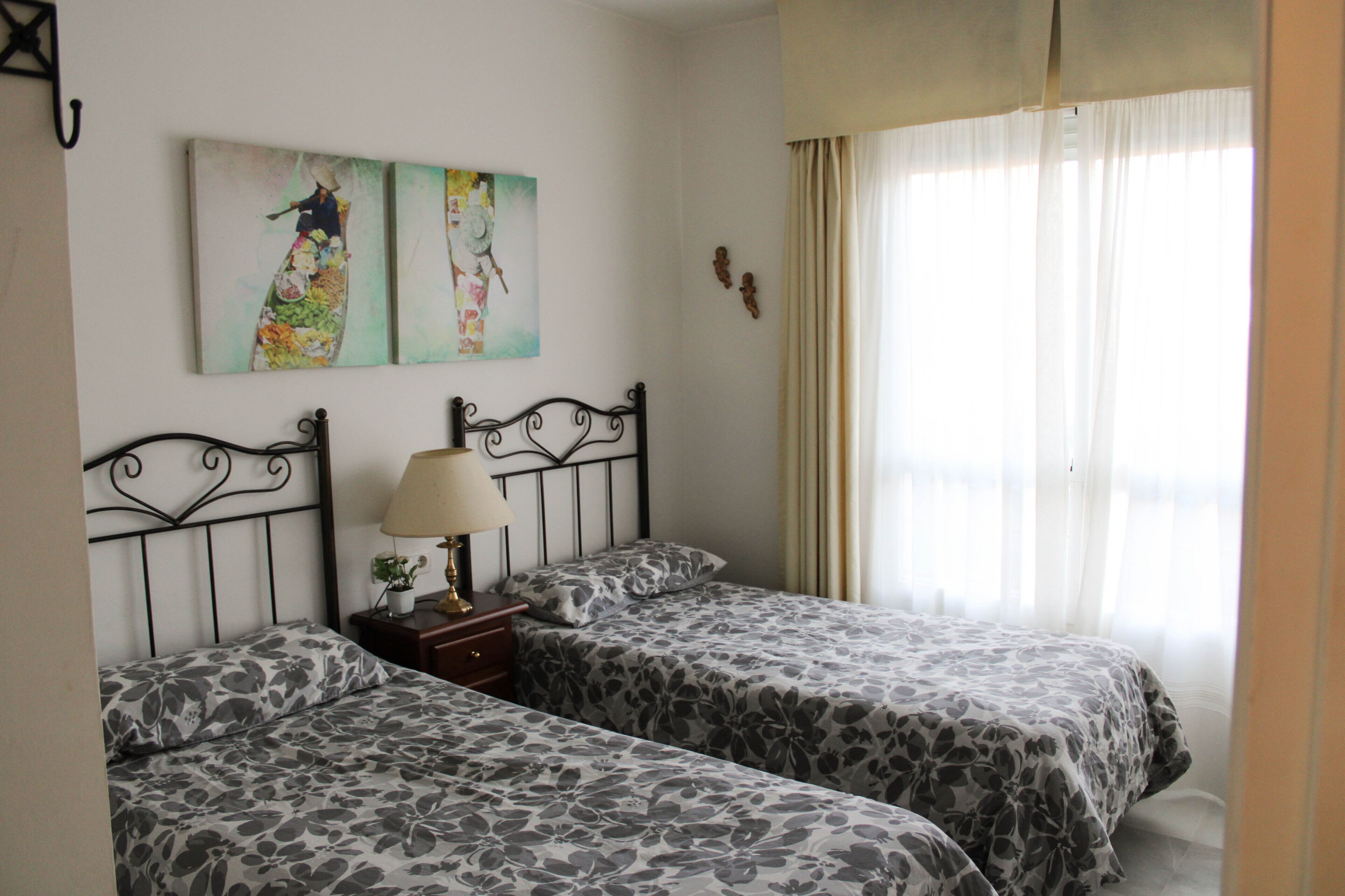 A room with two beds, a nightstand with a lamp, and two wall-mounted paintings.