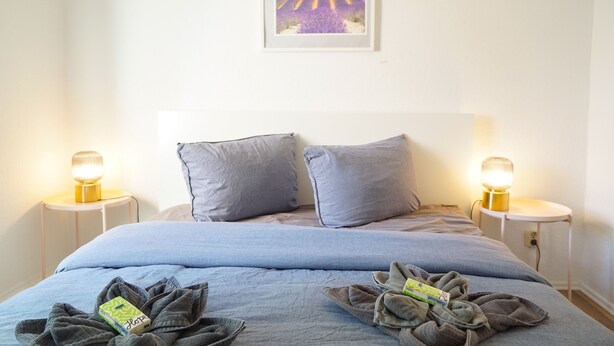 Een netjes opgemaakt bed met een blauwe deken en kussens, twee nachtlampjes en een lijst met een afbeelding van lavendelvelden aan de muur.