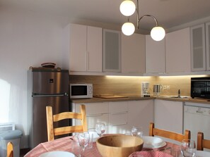 A compact kitchen with white cabinets, a stainless steel refrigerator, a microwave, and a coffee maker. There is a wooden dining table with chairs and a red and white checkered tablecloth. - Charming 1BR Apartment with Mountain View, Close to Slopes & Shops, Sleeps 6 (Vars)