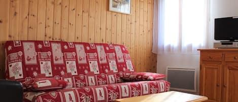 A cozy living room with a wooden paneled wall, a red patterned sofa, a wooden table, a television on a cabinet, and a window with curtains.