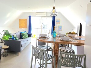 Una sala de estar moderna con un sofá gris, cojines azules y un comedor con una mesa de madera, sillas blancas y una botella azul.