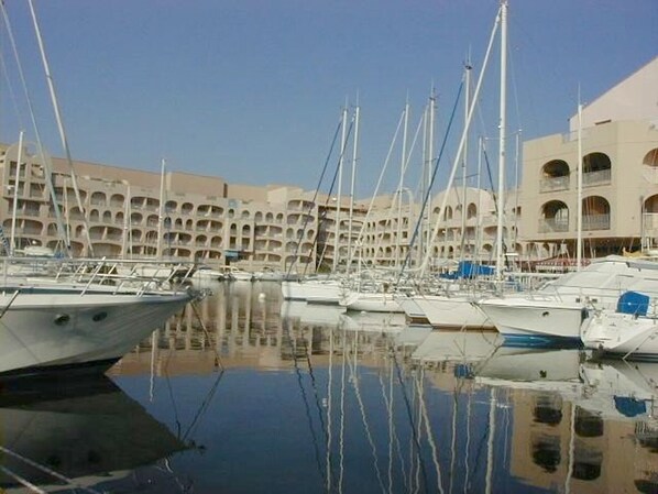 A marina with multiple boats docked, a large building with multiple balconies in the background, and clear skies.