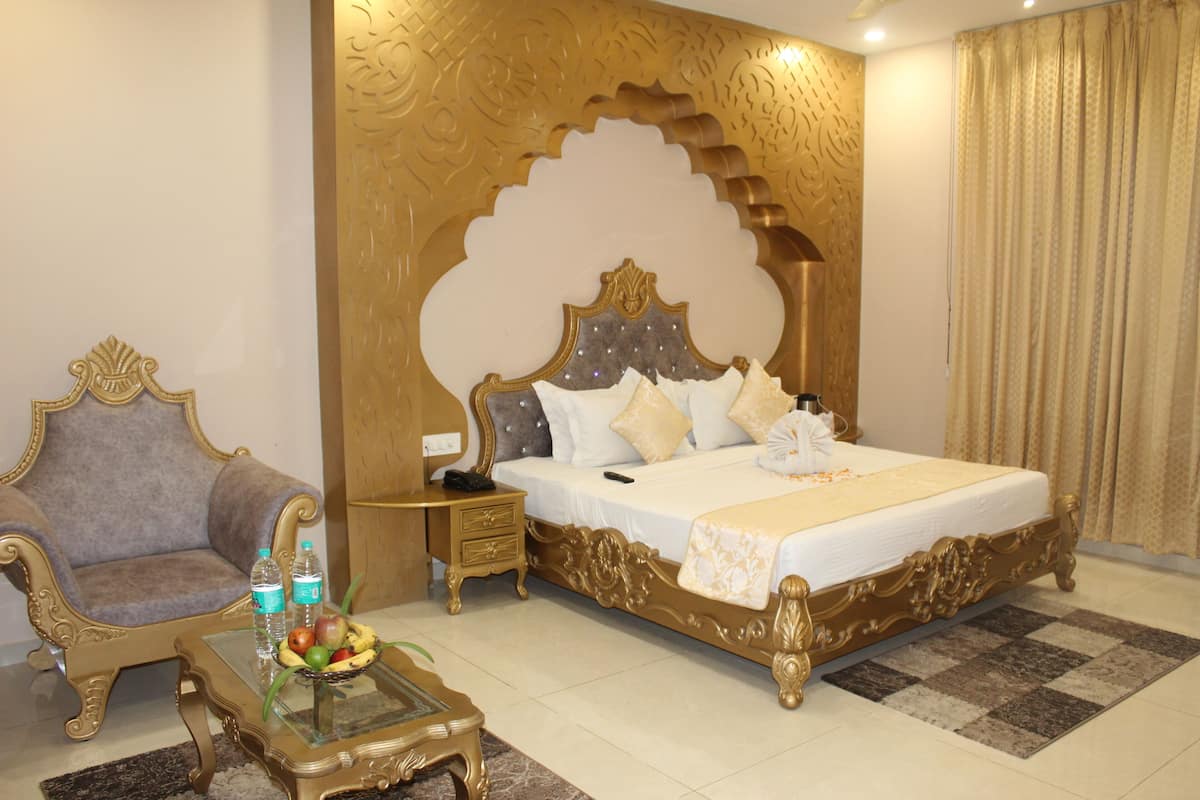 Quarto de hotel com cama grande, uma cadeira, mesinha com frutas e garrafas, e decoração na parede.