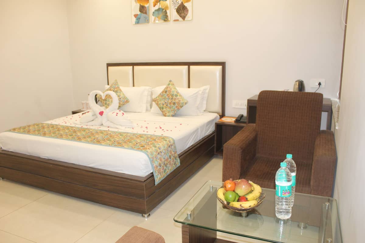 Quarto de hotel com cama, mesa de centro de vidro com frutas e uma garrafa de água, poltrona marrom e mesa de cabeceira com telefone e abajur.