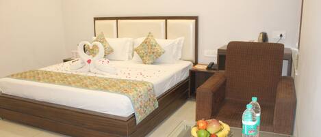 A hotel room with a bed, a glass coffee table with fruit and a water bottle, a brown armchair, and a nightstand with a phone and a lamp.