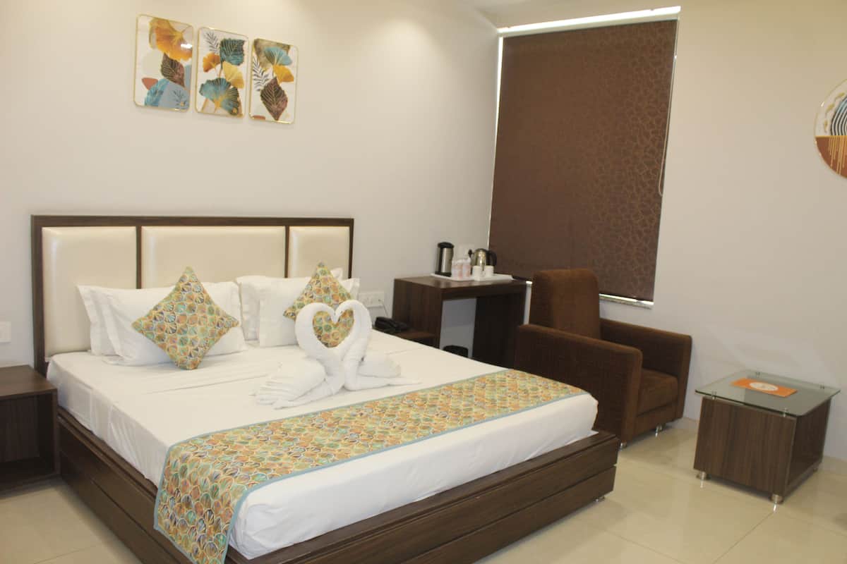 Quarto de hotel com uma cama grande, cabeceira de madeira, poltrona marrom, uma escrivaninha pequena com abajur e uma janela com persiana marrom.