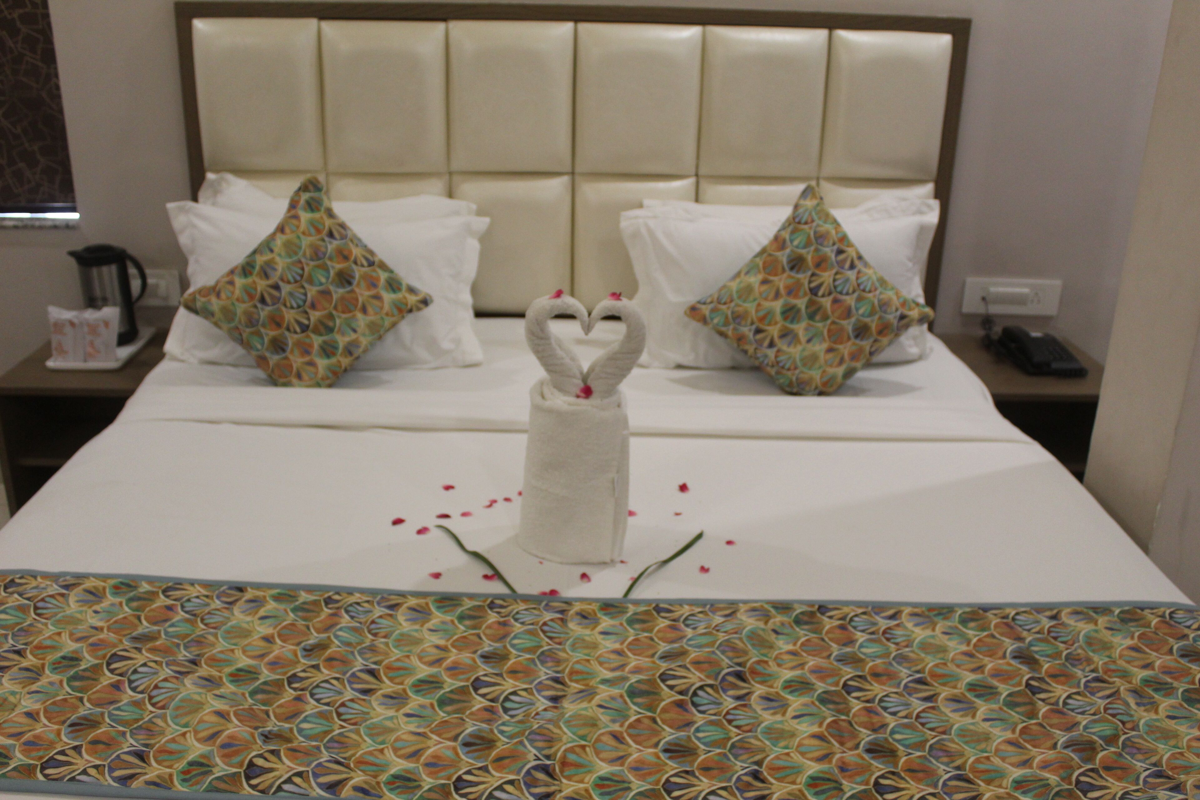 A hotel room with a bed featuring heart-shaped towels, a patterned bedspread, and a nightstand with a coffee maker and remote control.