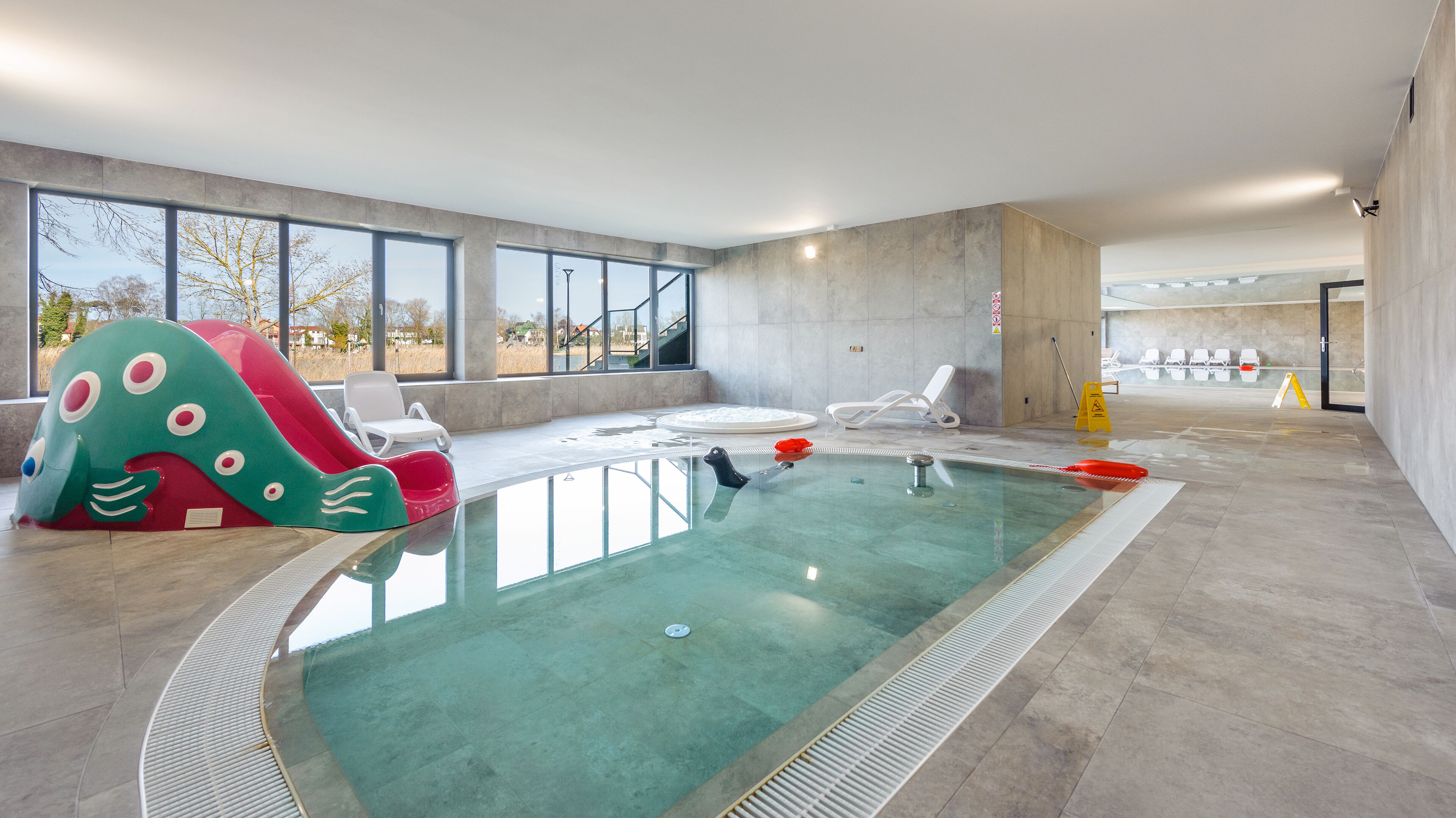 Piscina interna com um escorregador colorido, cadeiras de sol e vista para o exterior.