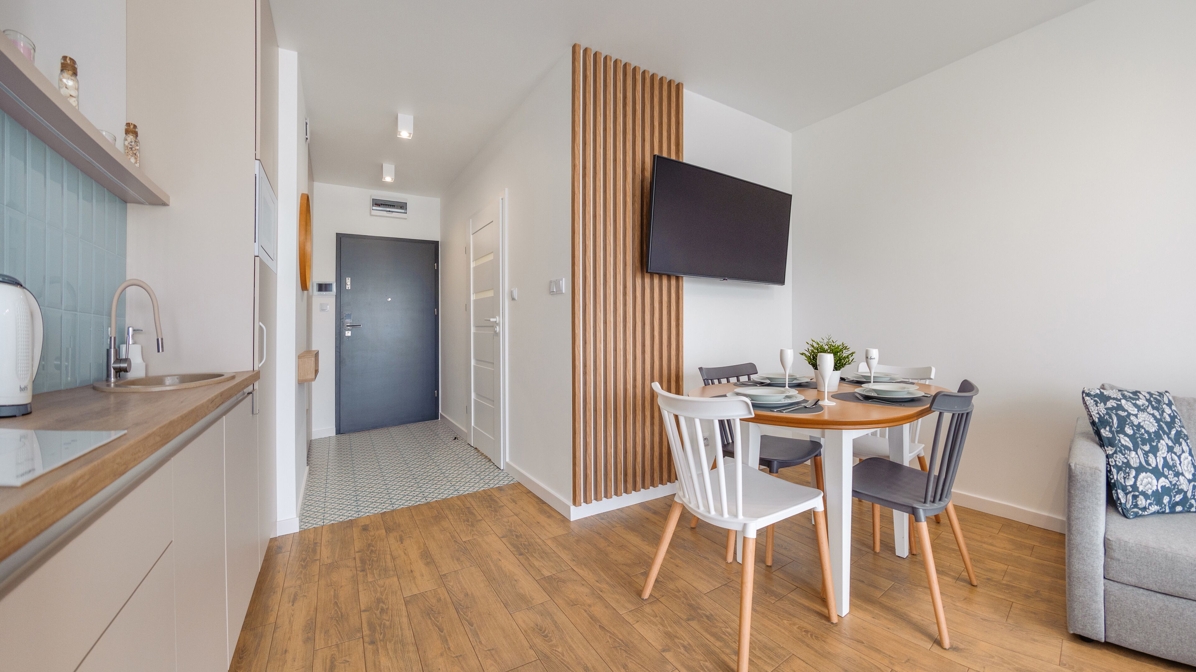 Ett modernt kök med en inbyggd spis, en diskho och en bänkskiva i trä. Här finns också en matplats med ett bord för två personer, en tv som är monterad på väggen och en soffa i hörnet.