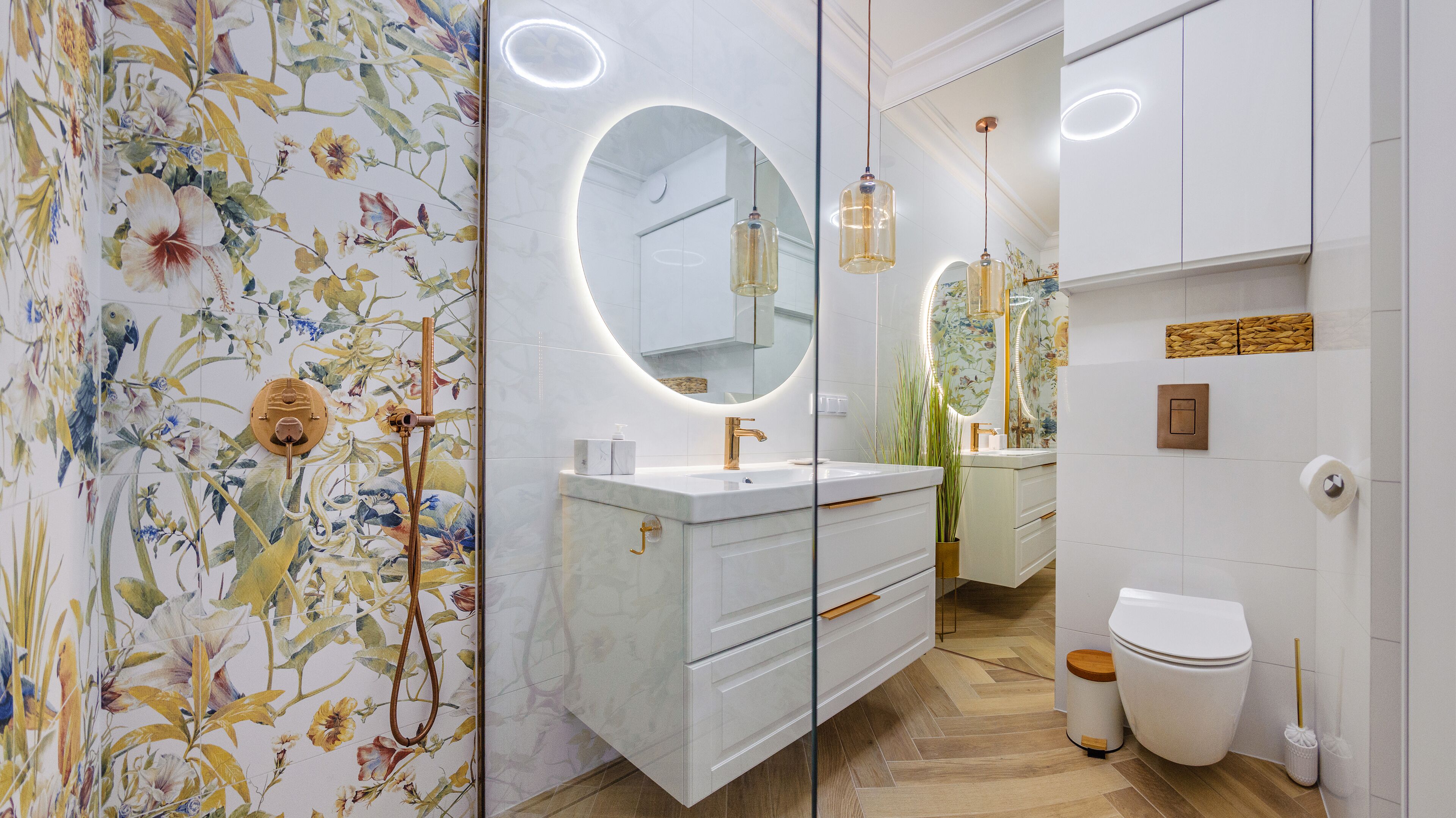 A modern bathroom with a floral wallpaper, a round mirror, a white vanity, and a toilet.