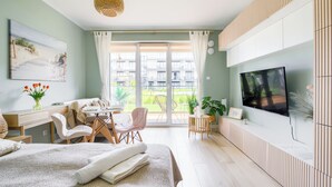 Quarto moderno com cama, escrivaninha, cadeira, televisão e varanda com vista para edifícios e vegetação.